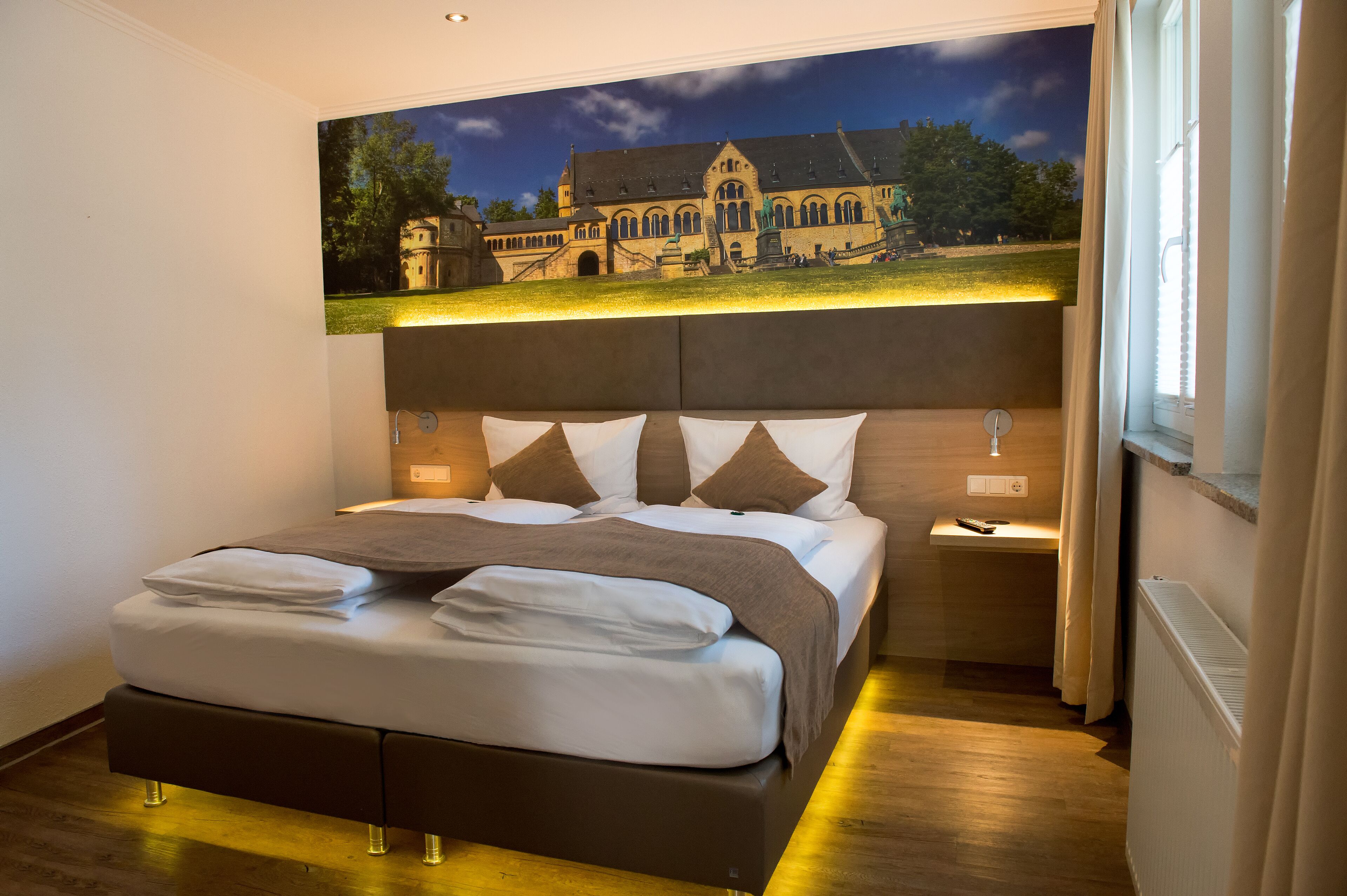 Ein modernes Hotelzimmer mit einem großen Bett, einem Nachttisch und Blick auf ein historisches Gebäude durch ein Fenster.