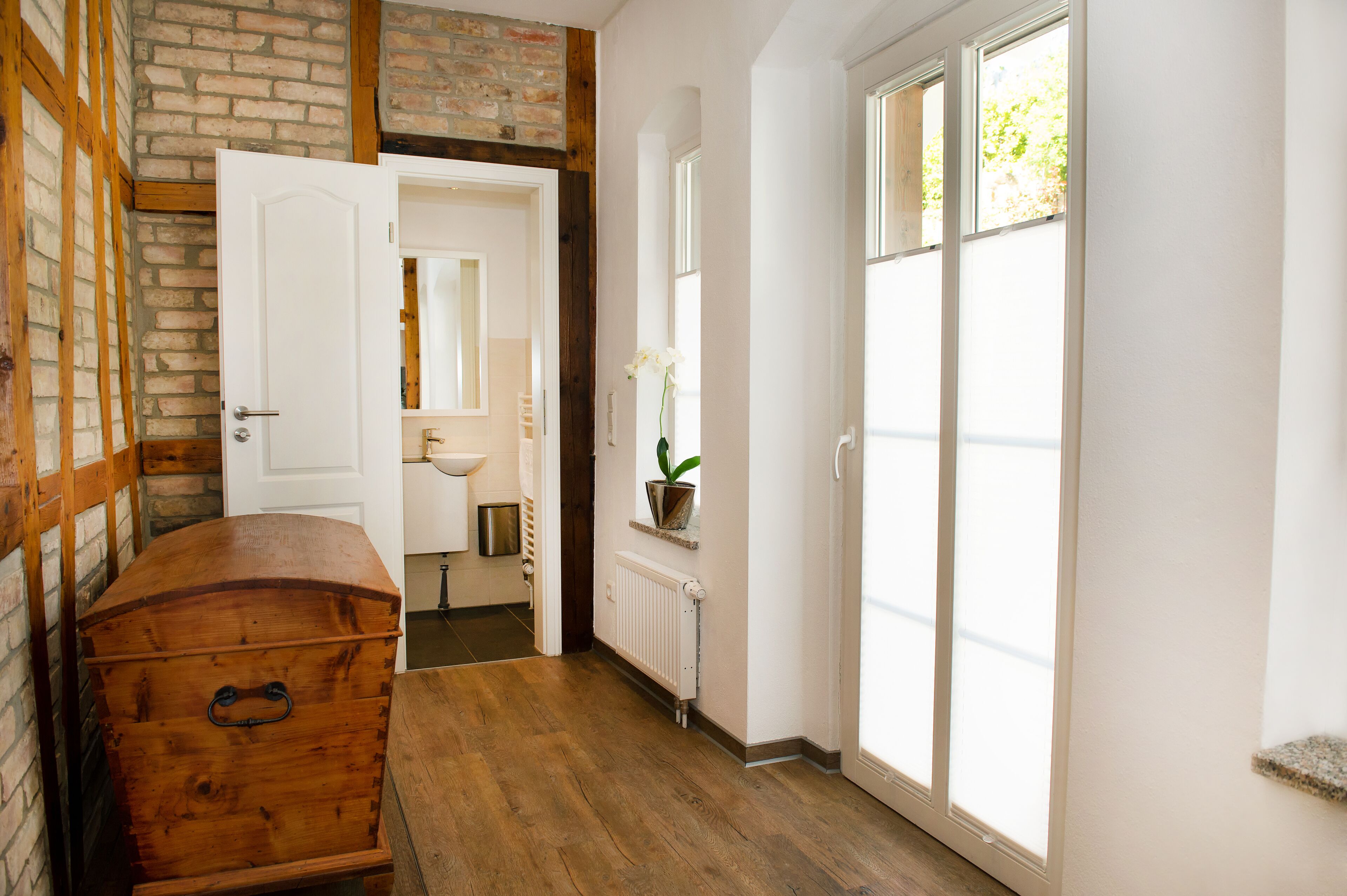 Un couloir avec une vieille malle en bois, une porte blanche donnant sur une salle de bain et une fenêtre d’où l’on peut voir les arbres.