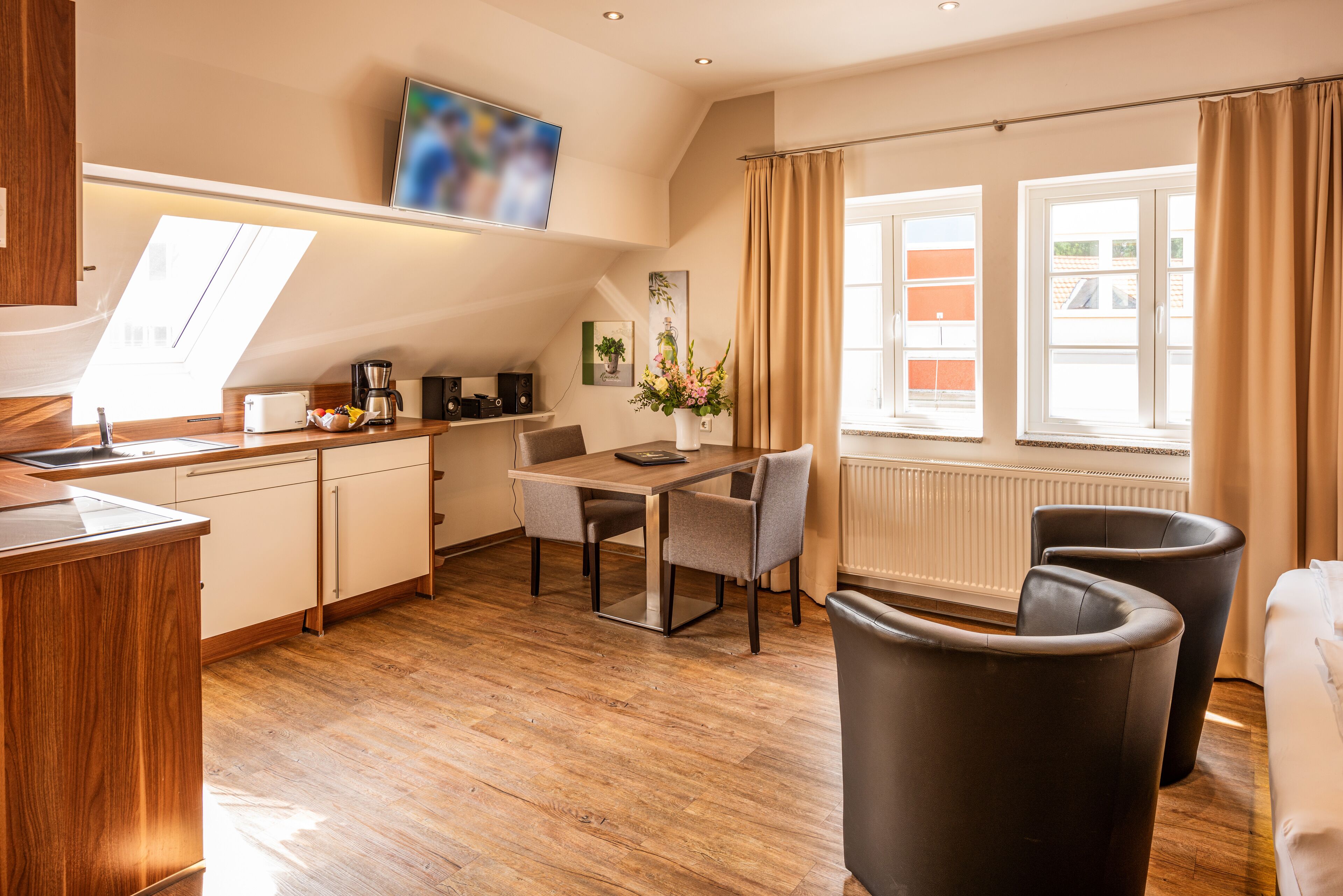 A modern kitchen with a dining area, wooden flooring, and a large window with curtains.