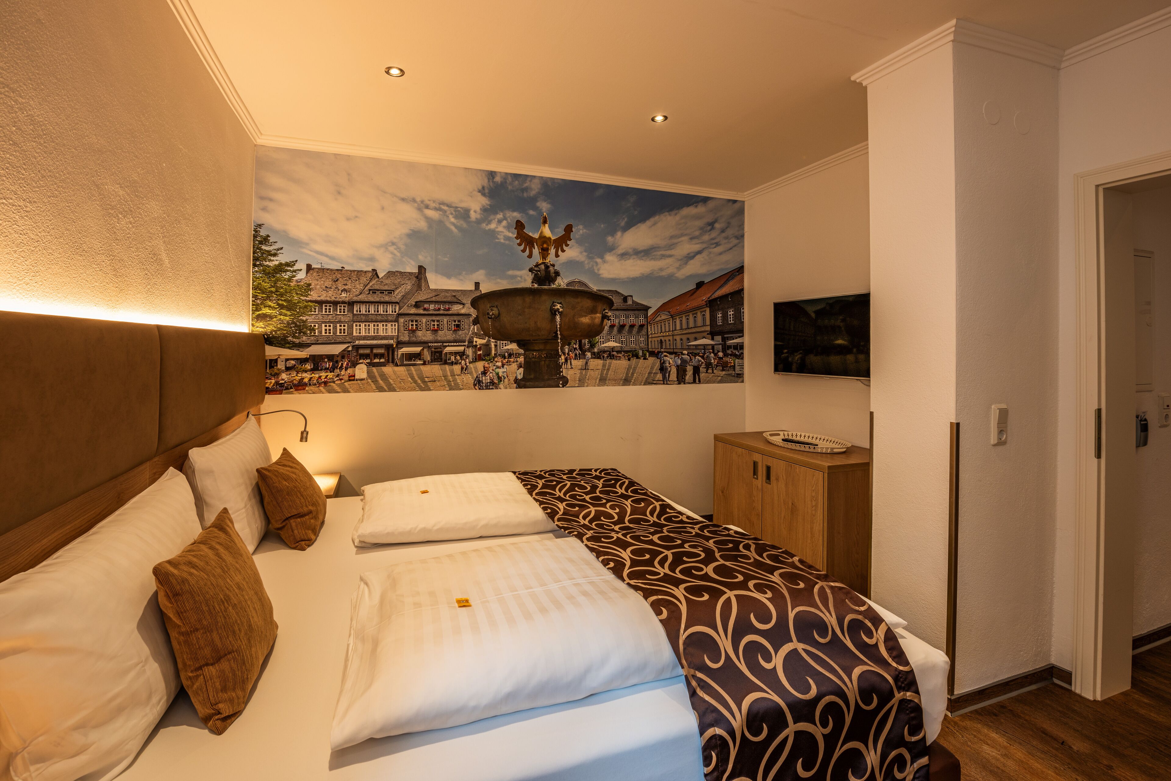 Een hotelkamer met een bed, een houten hoofdeinde, een muurschildering van een stadsplein en een klein kastje.