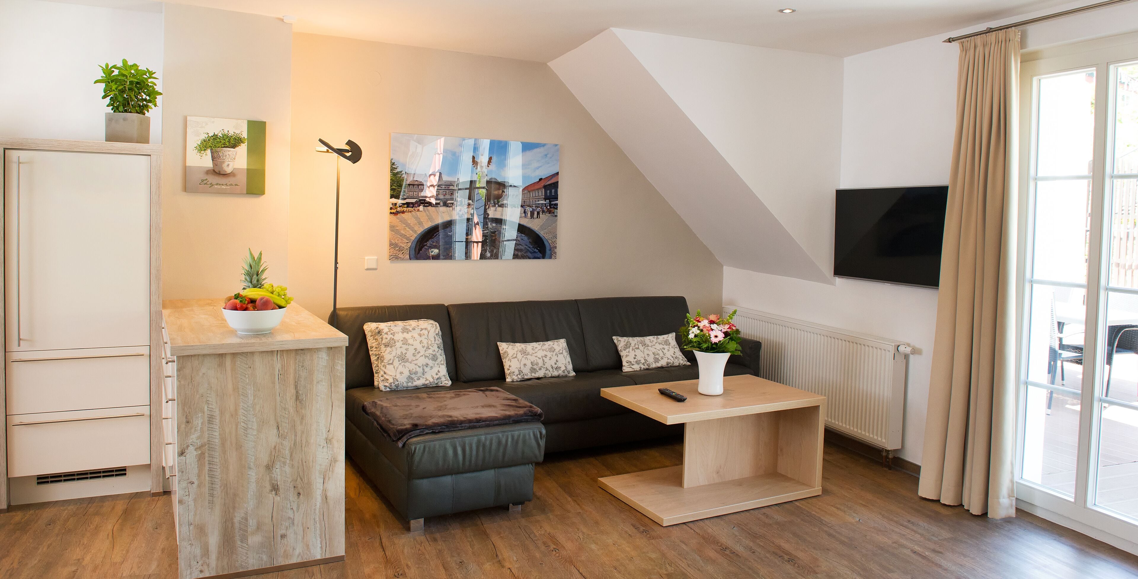 Een moderne woonkamer met een bank, salontafel, televisie en een keukenhoek met een koelkast en een plant.