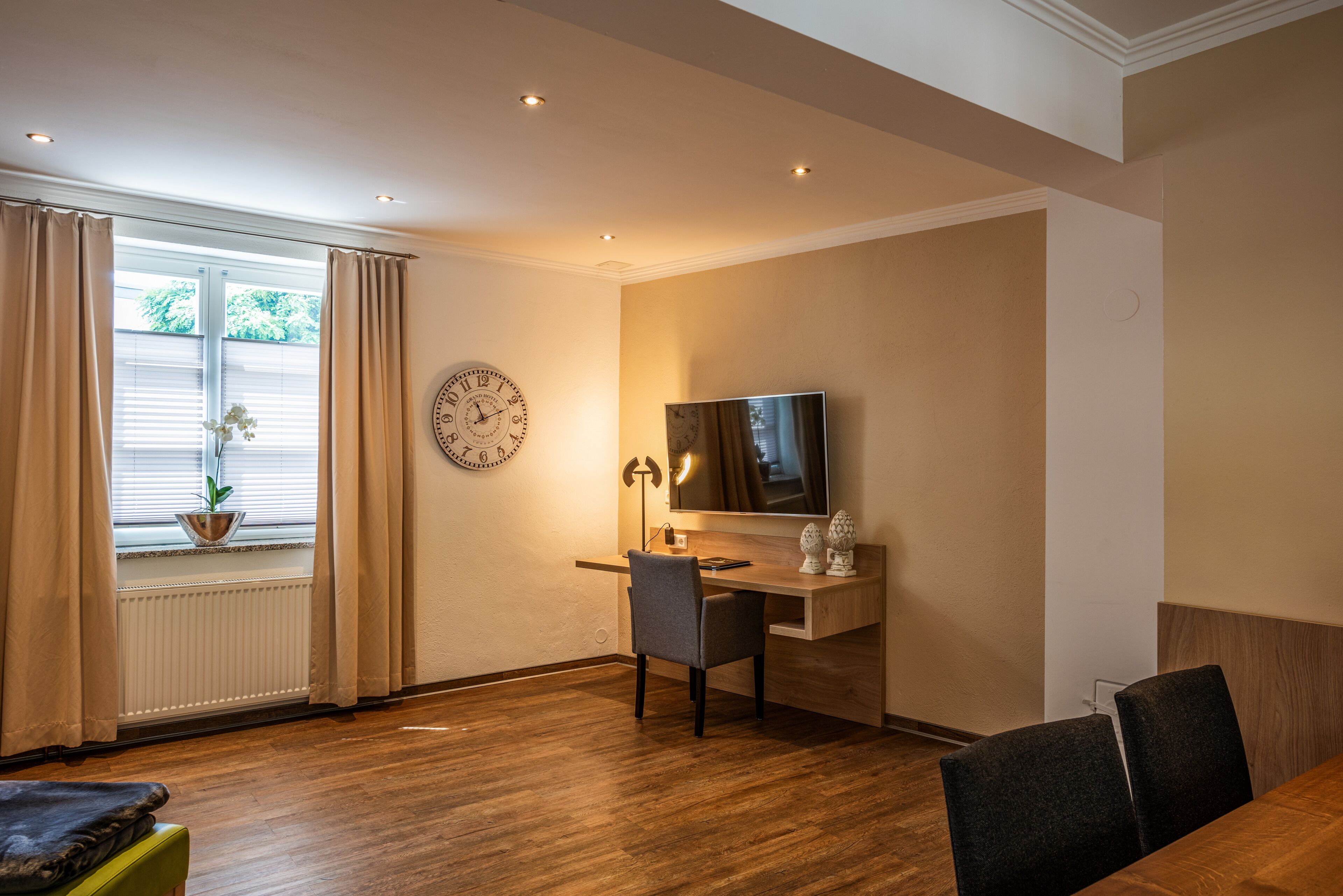 Una sala de estar moderna con piso de madera, un escritorio con una silla, un televisor, un reloj y una ventana con cortinas.