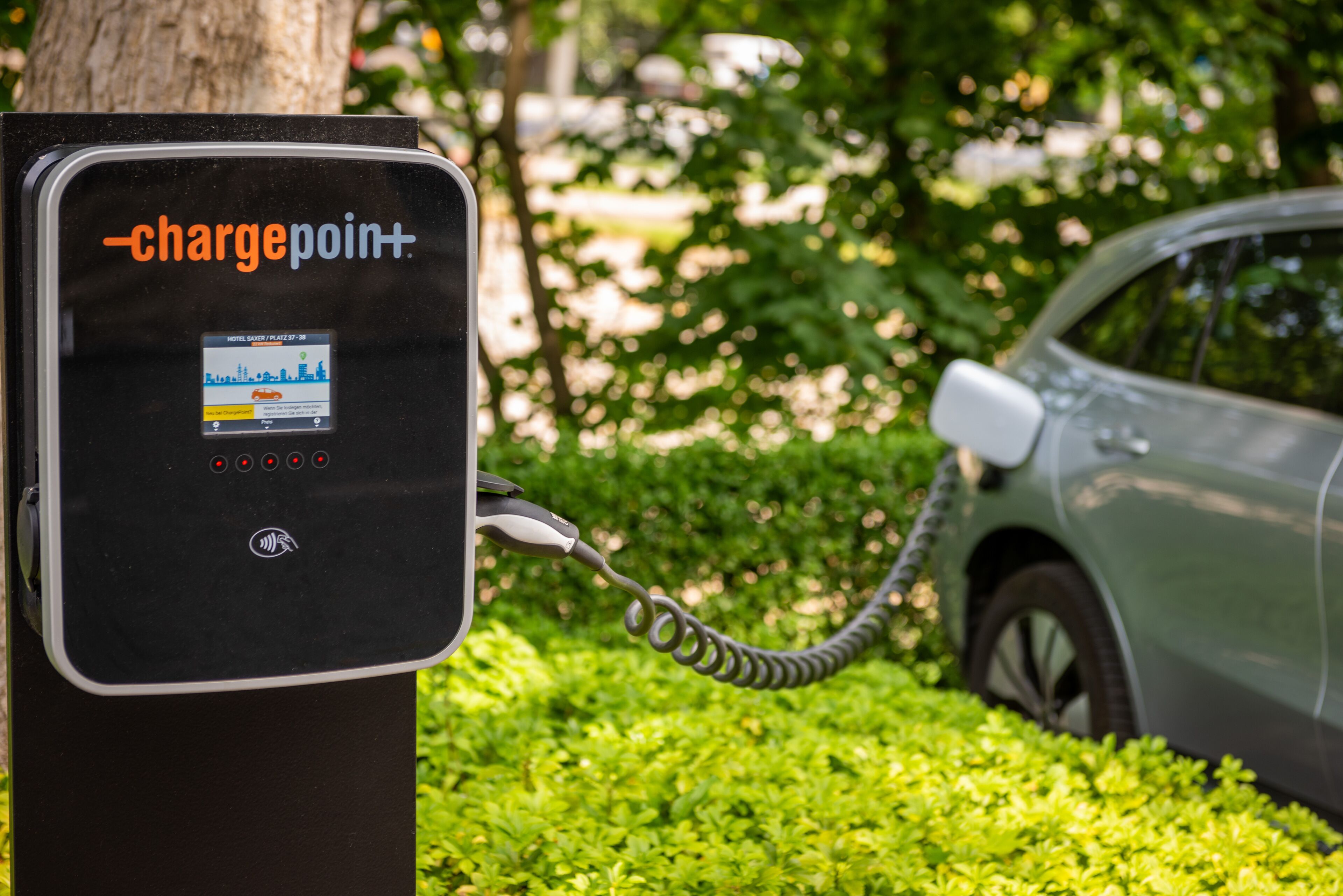 A chargepoint+ charging station with a car connected to it, set against a backdrop of greenery.