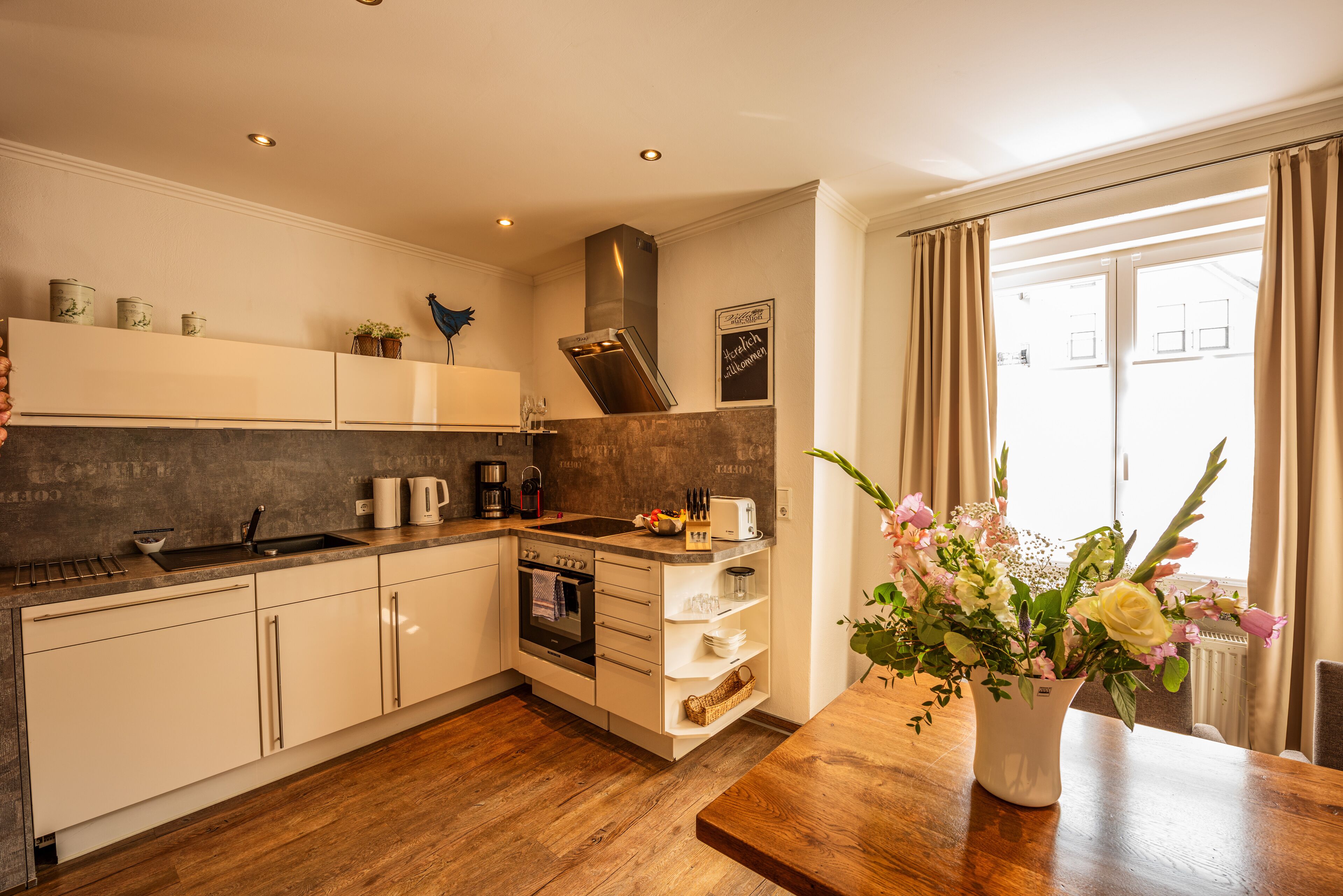 A modern kitchen with white cabinets, a wooden countertop, and a built-in oven. There is a window with curtains and a vase of flowers on the counter.