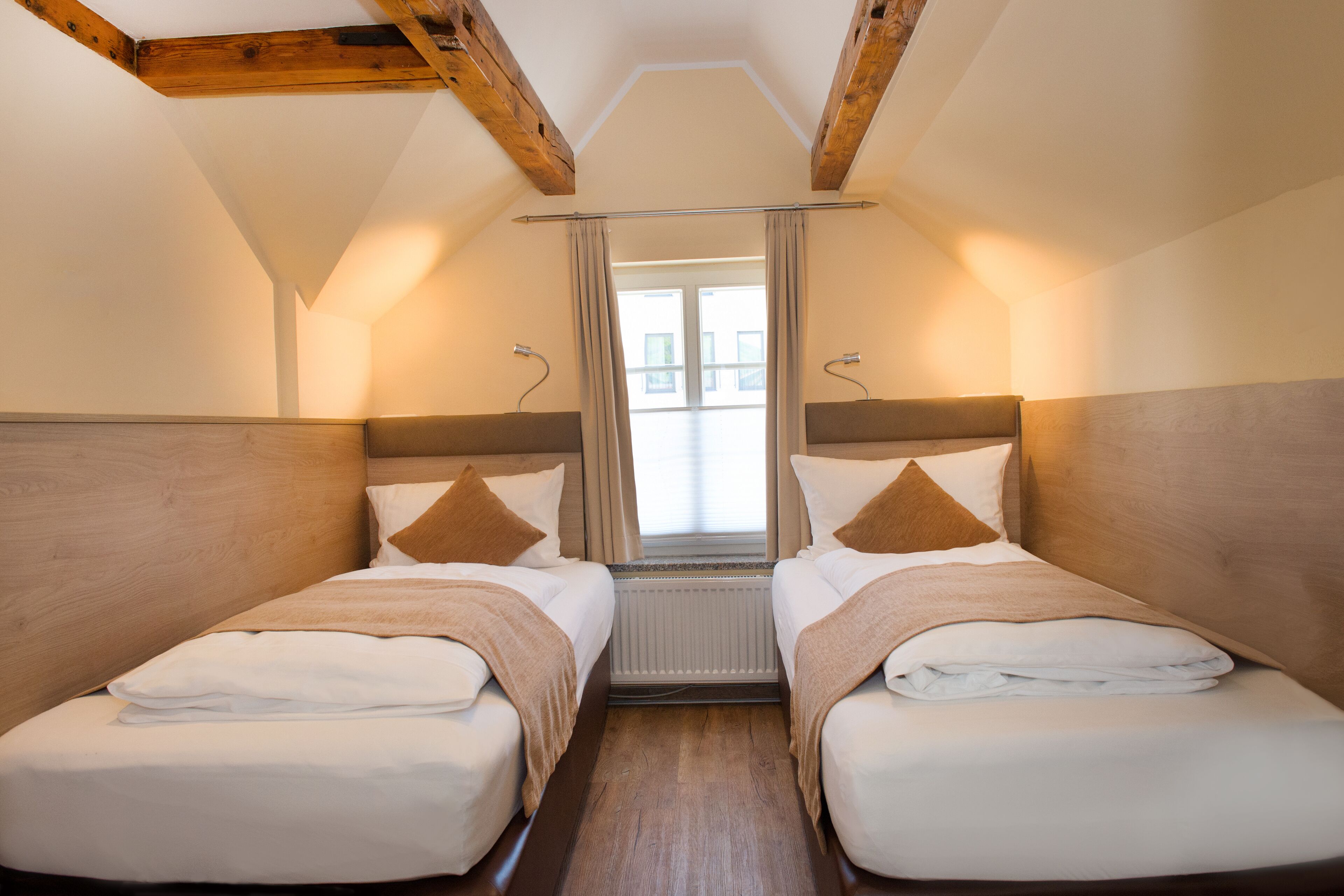 Ein Zimmer mit zwei Betten, Holz-Kopfteilen, einem Vorhangfenster und einem Heizkörper zwischen den Betten.