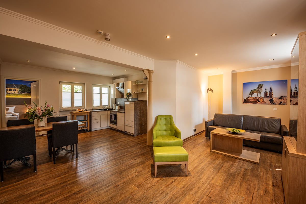 Een ruime woonkamer met een houten vloer, een groene fauteuil, een bank en een eettafel met stoelen.