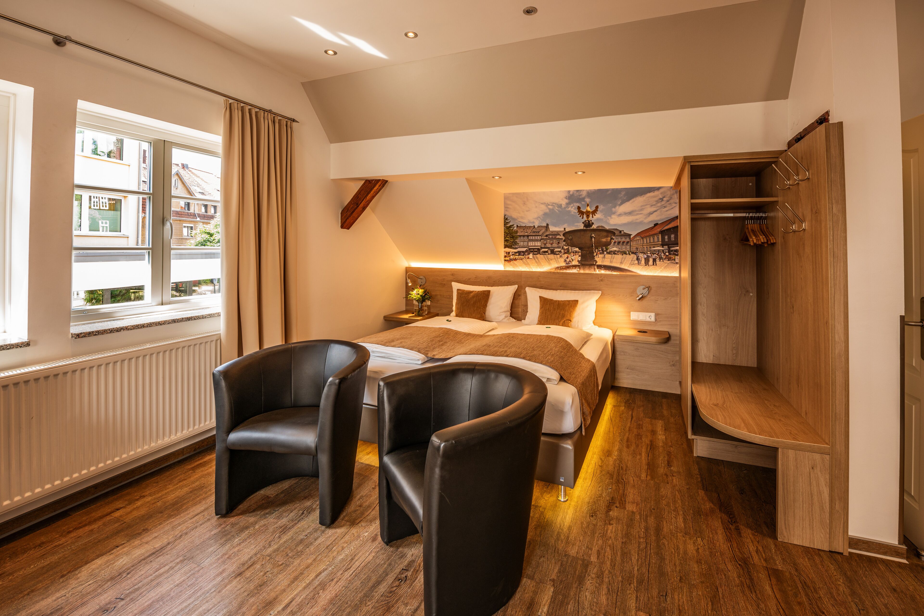 A modern hotel room with a large bed, two black leather chairs, a wooden desk, and a view of a town through a window.