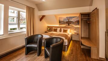A modern hotel room with a large bed, two black leather chairs, a wooden desk, and a view of a town through a window.
