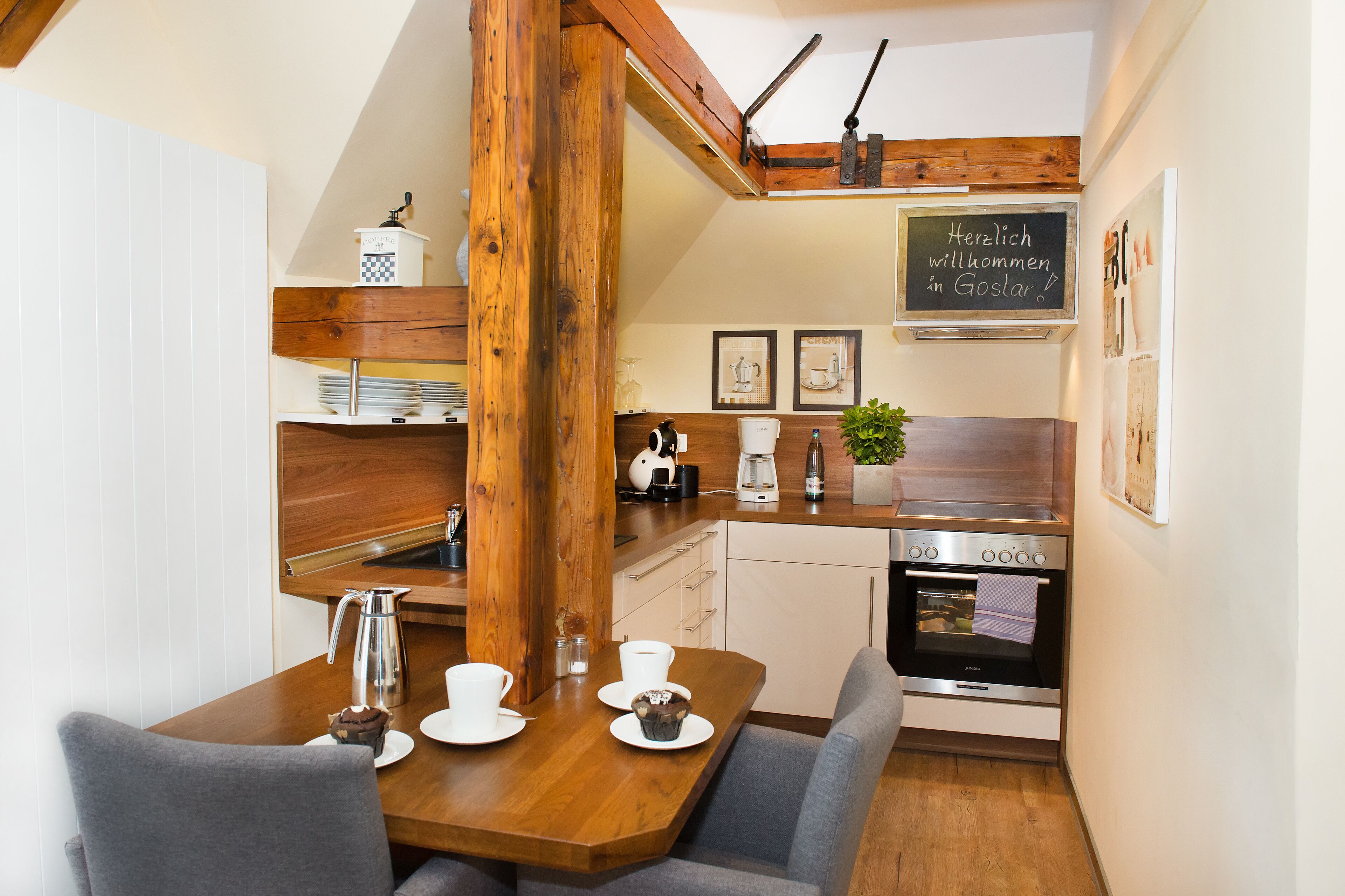 A compact kitchen with wooden beams, a coffee maker, a kettle, and a chalkboard with a welcome message.