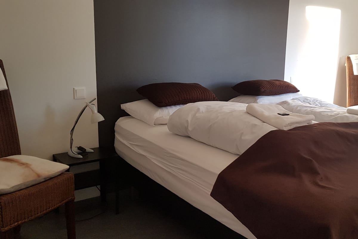 A neatly made bed with white and brown bedding, a bedside table with a lamp, and a chair with a cushion.