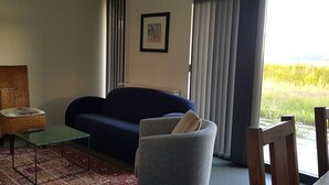 Ett vardagsrum med en blå soffa, en grå fåtölj, en flätad stol, ett litet bord och utsikt över ett fält genom fönstret.