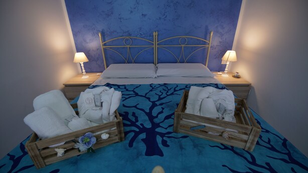 A hotel room with a bed, two bedside tables with lamps, a blue patterned bedspread, and two wooden cradles with towels. - B&B Nodo Francescano (Porto Empedocle)