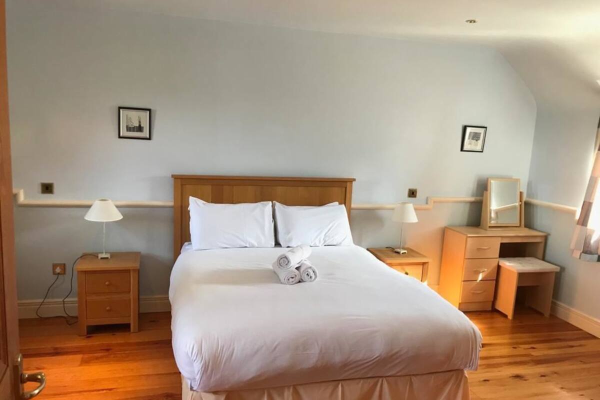 A neatly made bed with white linens and rolled towels, wooden headboard, bedside tables with lamps, and a small desk with a mirror.
