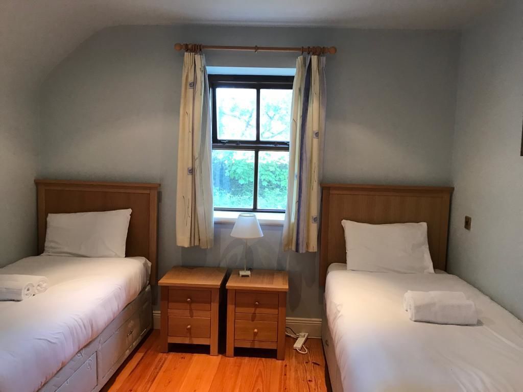 Quarto com duas camas de solteiro, uma mesa de cabeceira de madeira com gavetas, uma luminária e uma janela com cortinas.