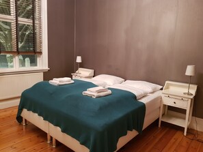 A neatly made bed with a teal blanket, white pillows, and two folded towels on the side. There is a bedside table with a lamp and a small drawer. The room has wooden flooring and a window with blinds. - Casa Apartments (Reykjavik)