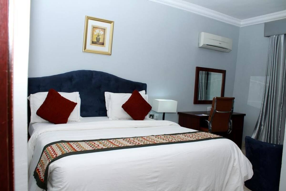A hotel room with a bed, a desk, a chair, a lamp, an air conditioning unit, and a framed picture on the wall.