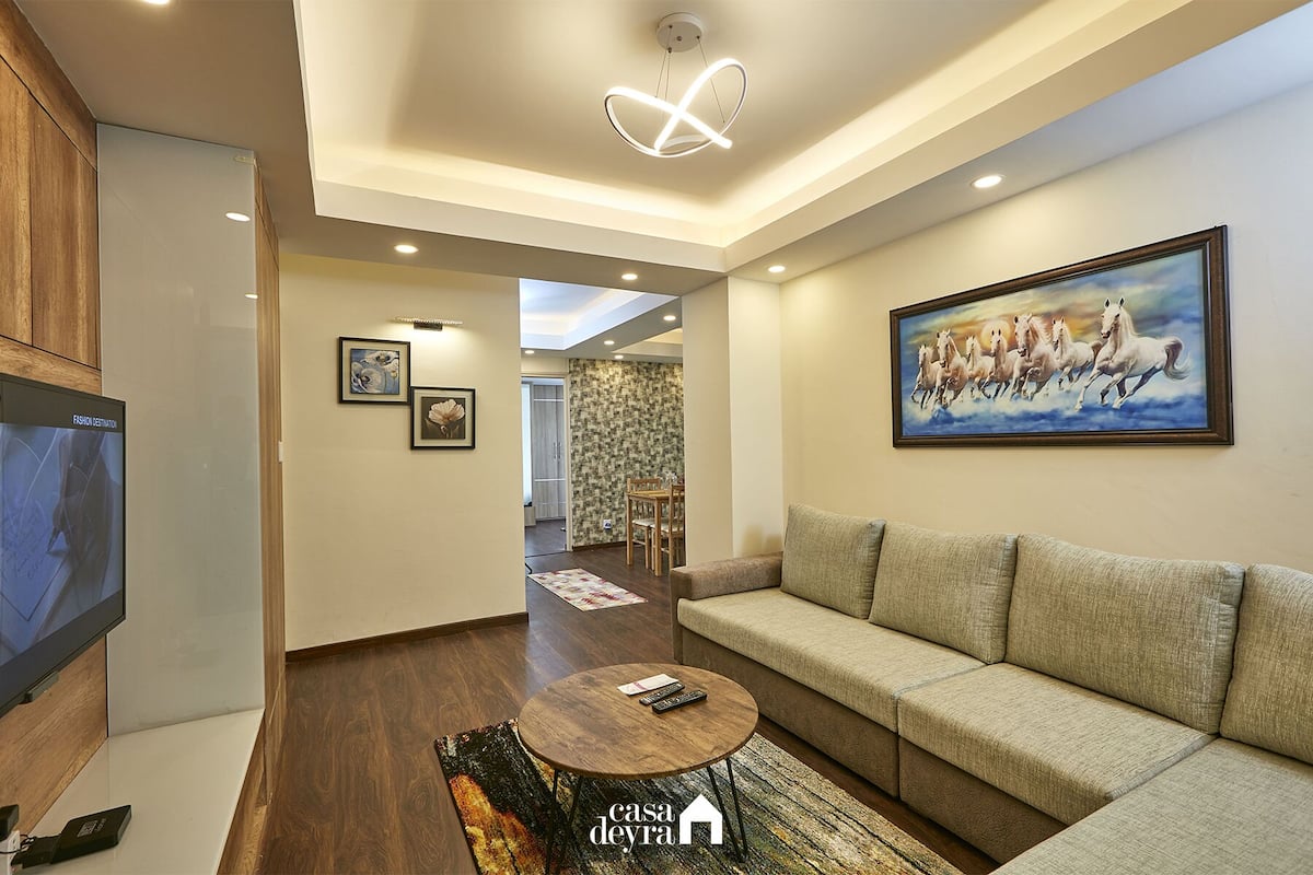 A modern living room with a sofa, a coffee table, and a TV. There is a painting on the wall and a ceiling light fixture.