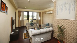 A living room with a sofa, a coffee table, and a TV. There are curtains, a window, and a wall-mounted air conditioner.