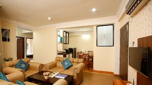 A living room with a sofa set, a coffee table, and a TV. There is a dining area in the background.