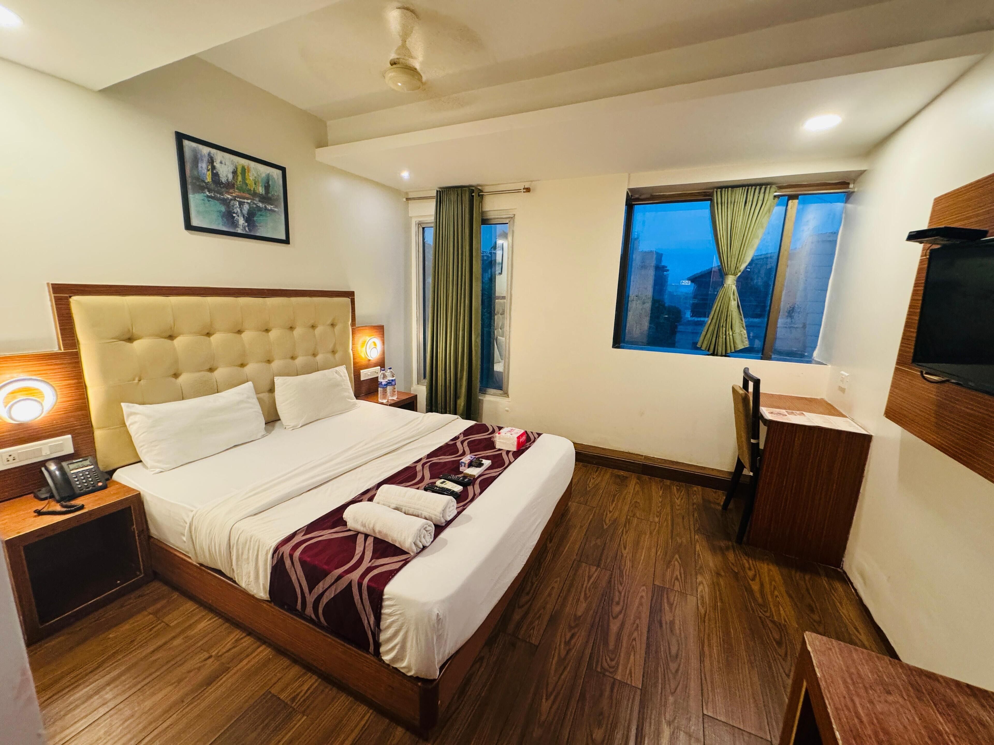 A hotel room with a large bed, bedside tables, a desk, a chair, a TV, and a window with curtains.
