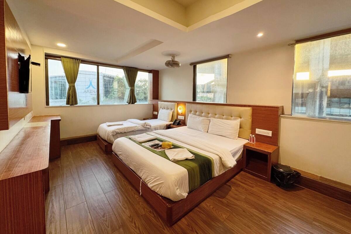 A hotel room with two beds, wooden flooring, a desk, and large windows with curtains.