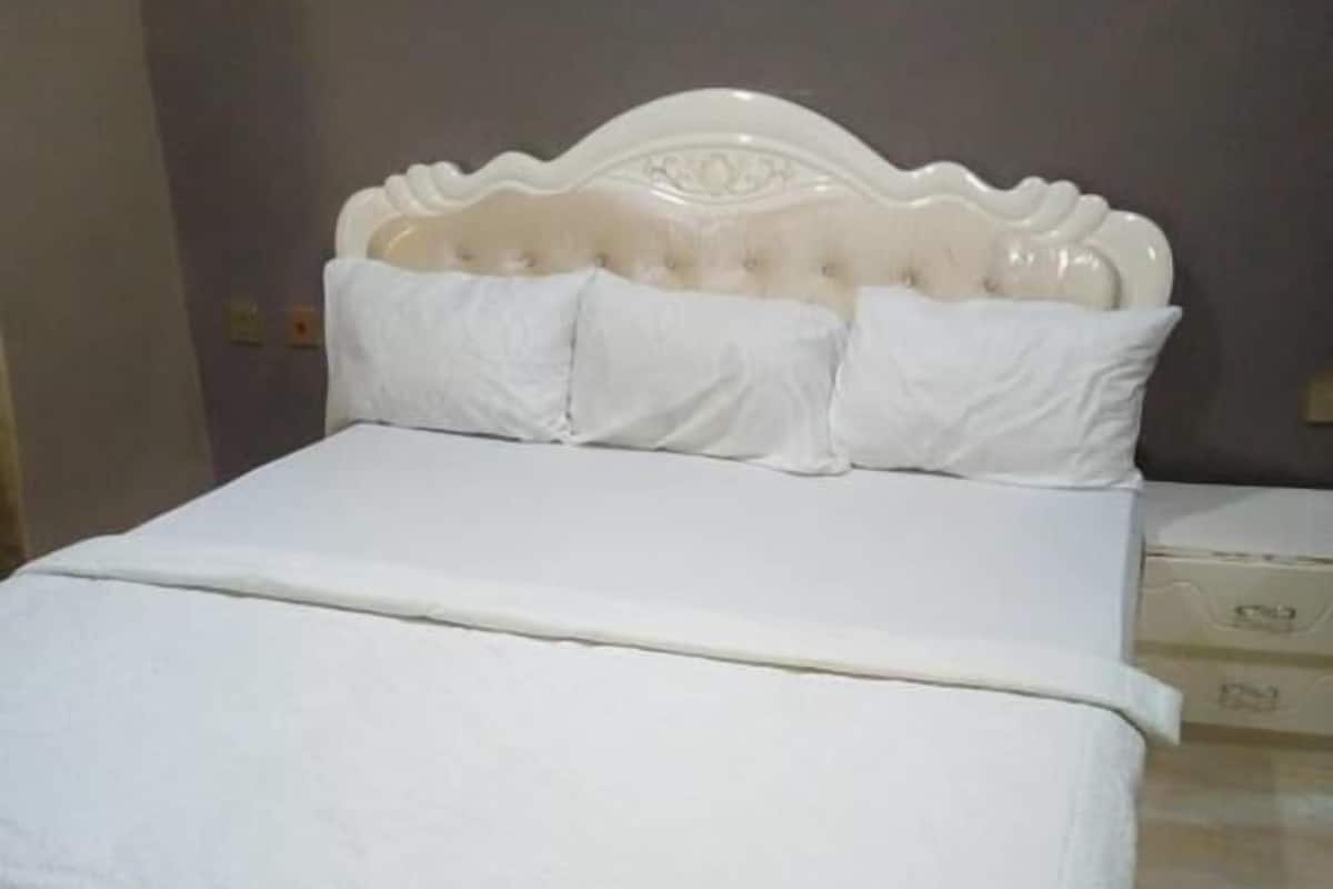 Una cama con cabecera blanca y almohadas, una colcha blanca y suelo de baldosas.