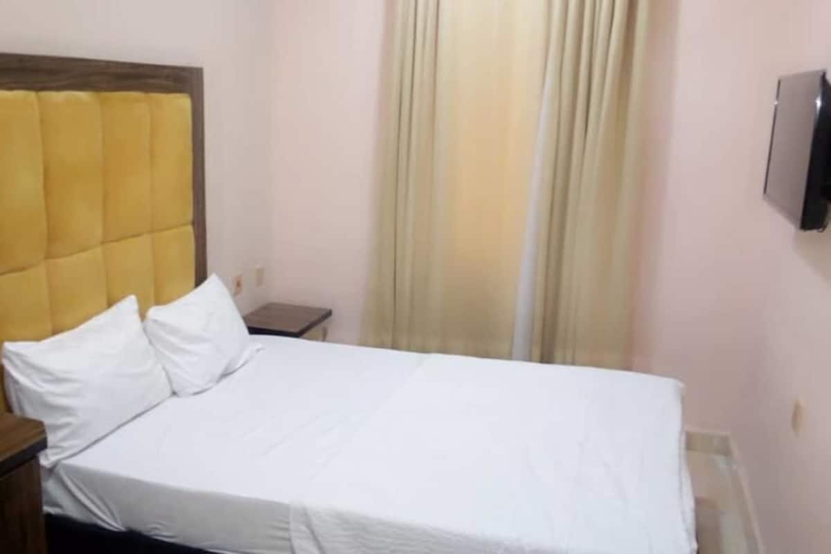 Una cama individual con cabecera de madera, una mesita de noche, un televisor montado en la pared y una ventana con cortinas.