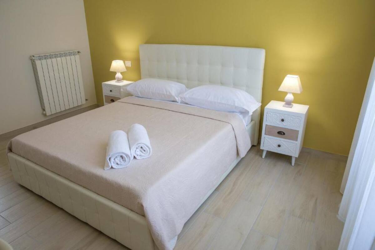 Cama bem arrumada com lençóis brancos e duas toalhas enroladas, cabeceira branca, duas mesas de cabeceira com luminárias e um radiador na parede.