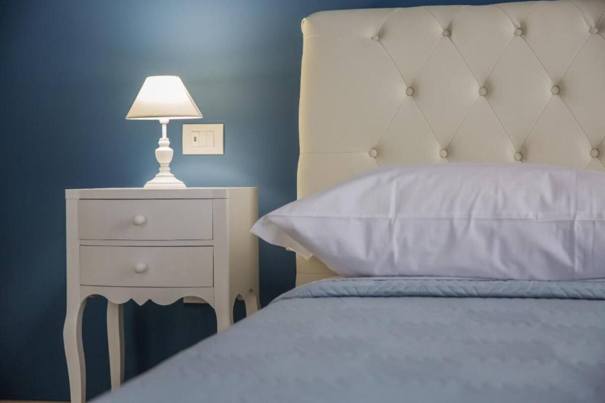 Quarto com mesa de cabeceira branca, luminária, cabeceira estofada e cama com roupa de cama branca.