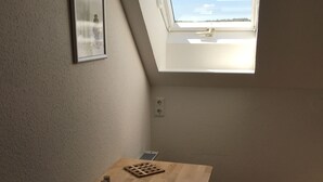 Une petite pièce avec une lucarne, une table en bois, une chaise et un tableau encadré au mur.