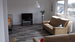 Un salon avec un téléviseur posé sur un meuble, un canapé, un fauteuil, une plante en pot et une fenêtre donnant sur des immeubles.