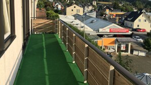 Un balcon doté d’un revêtement de sol synthétique vert, donnant sur une ville où l’on peut apercevoir différents bâtiments, notamment une station-service et une enseigne pour Terrassendächer.