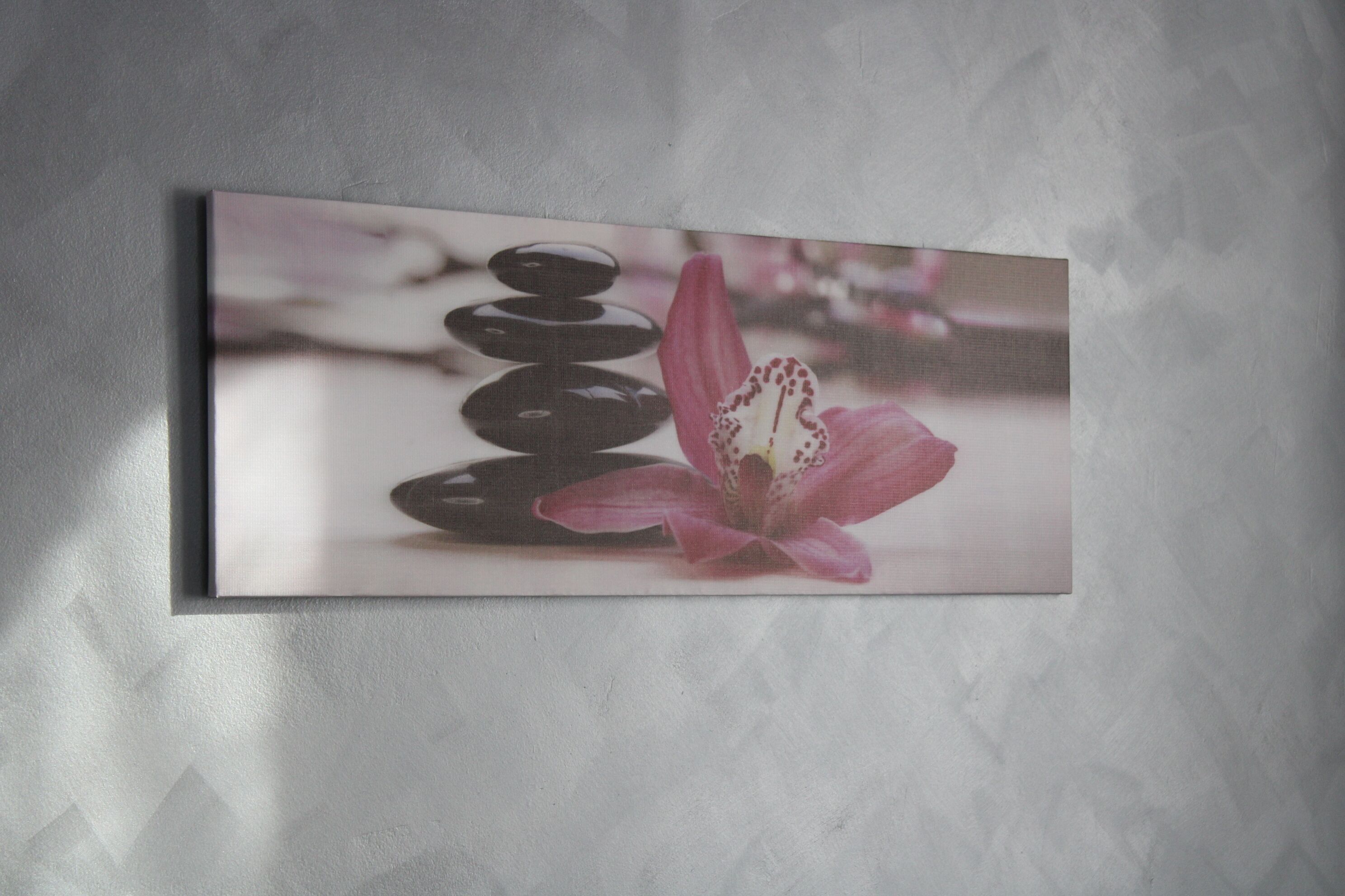 A framed artwork of stacked stones and a pink flower on a textured wall.