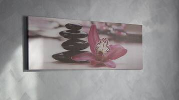 A framed artwork of stacked stones and a pink flower on a textured wall.