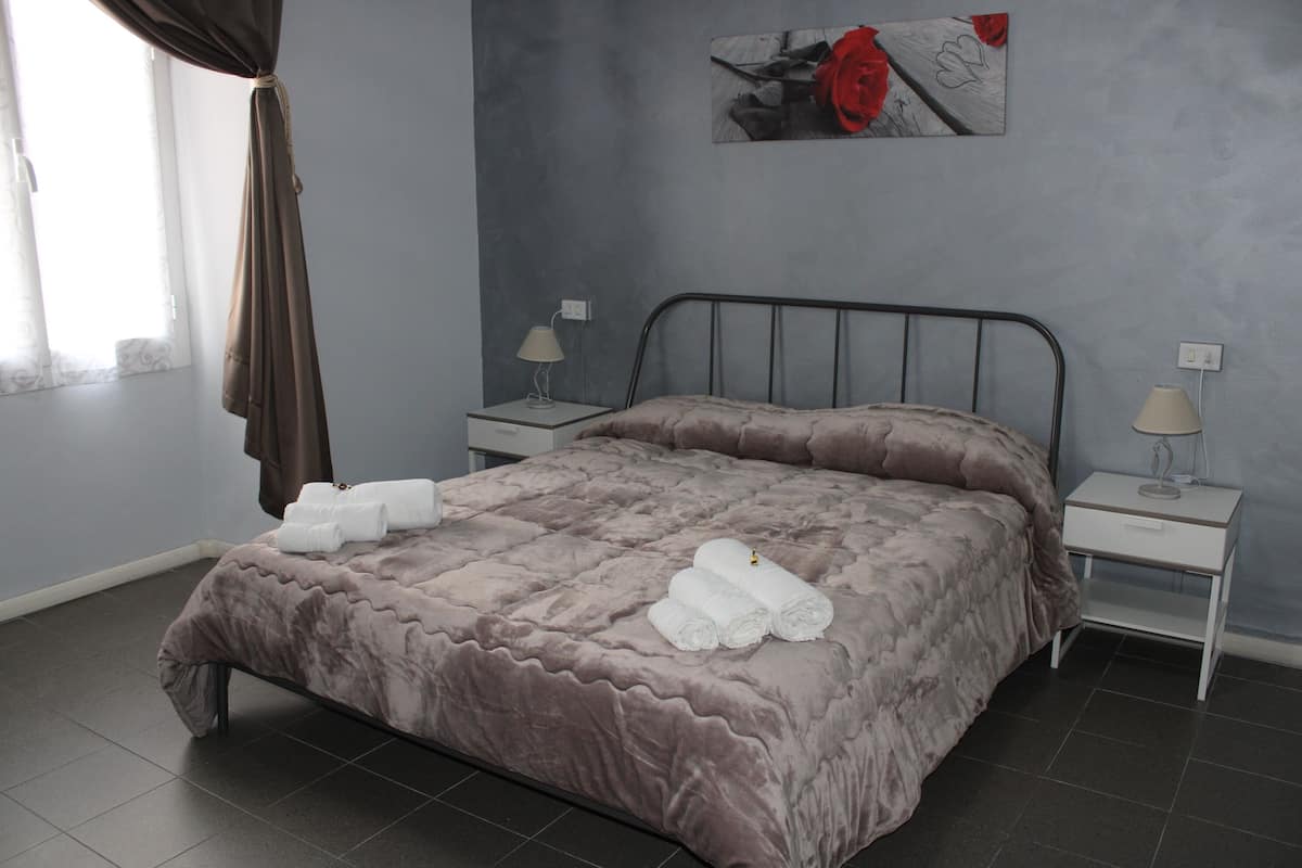 A neatly made bed with a plush, textured bedspread and rolled towels, two bedside tables with lamps, a wall-mounted artwork, and a window with curtains.