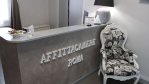Una reception con la scritta "AFFITTACAMERE ROMA", una poltrona decorativa dal design vintage, un calendario e una lampada.