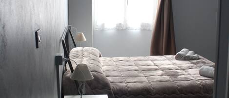 A bedroom with a bed, a desk with a lamp, a window with curtains, and a wall-mounted air conditioner.