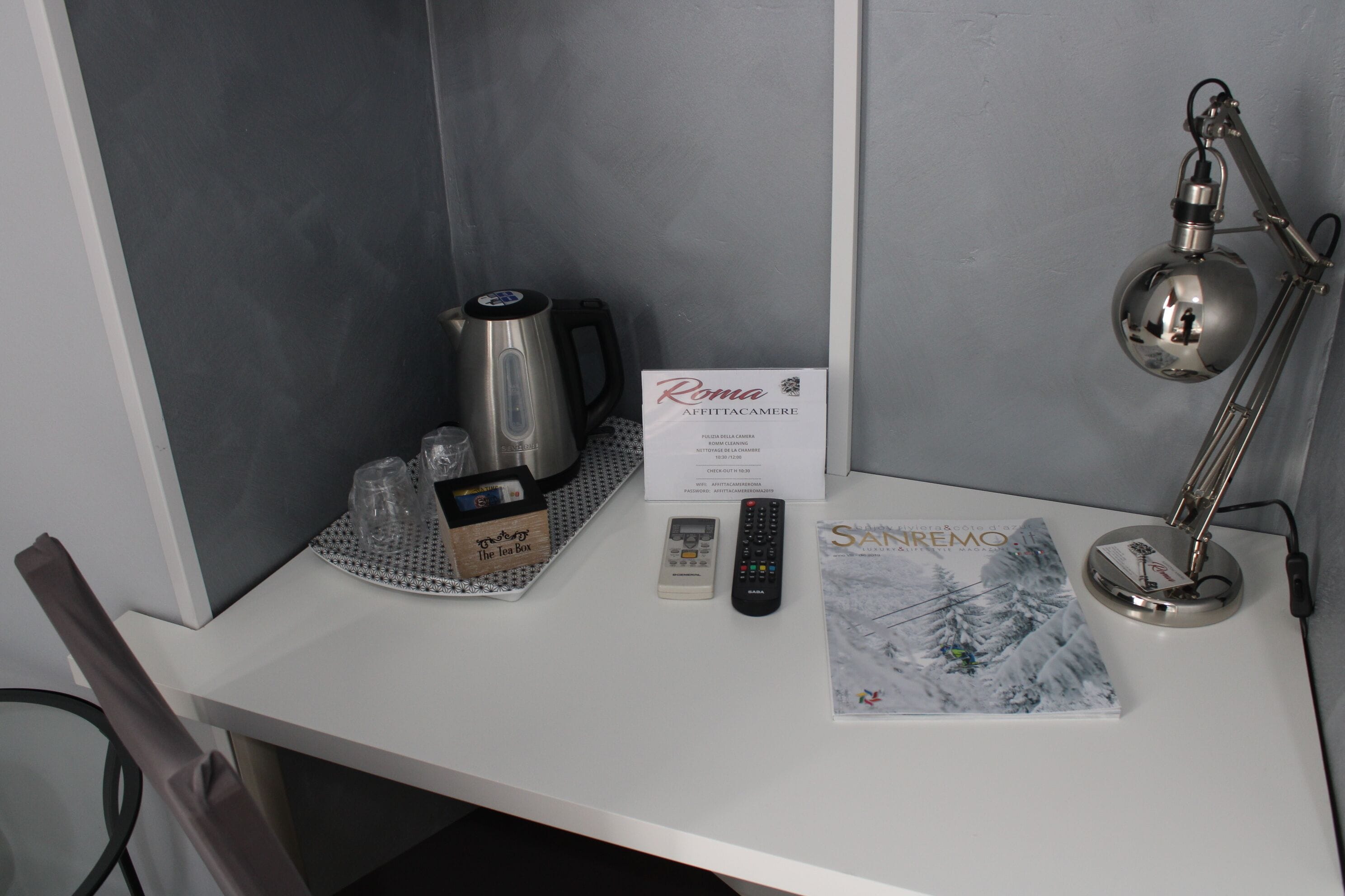 A desk with a kettle, a box labeled "The Tea Box," a remote control, a book titled "Sandro," and a lamp.