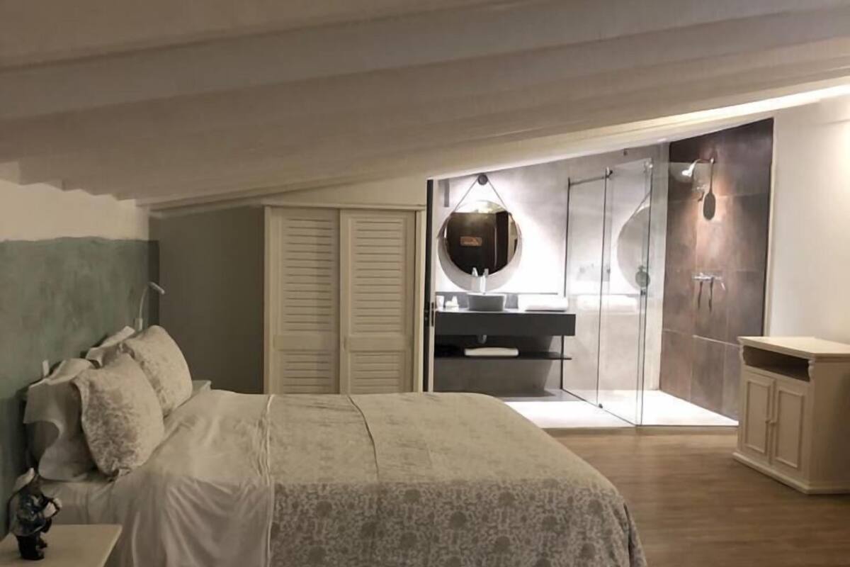 A bedroom with a bed, a nightstand, a dresser, and a bathroom with a sink and mirror.