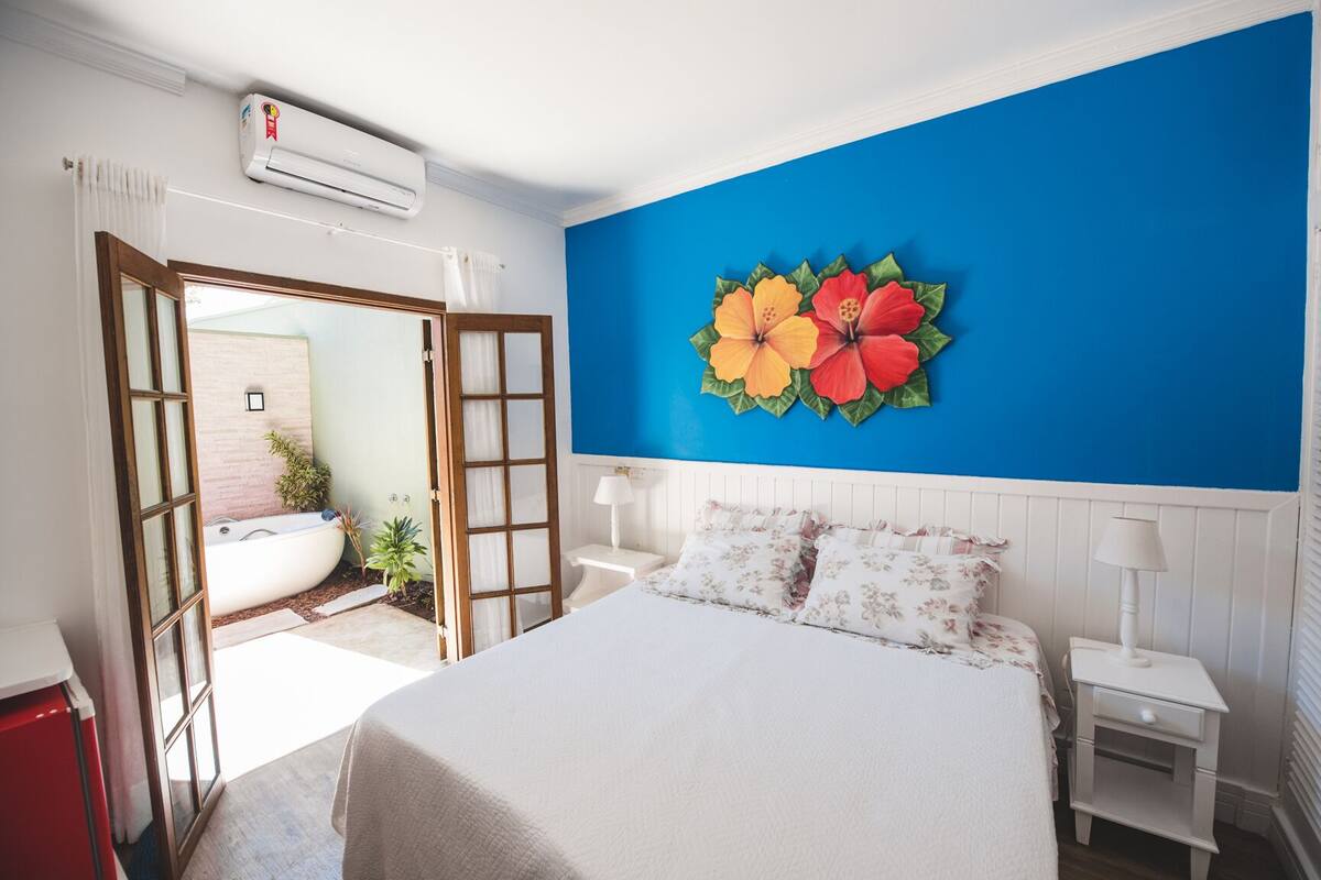 A bedroom with a bed, nightstands, a wall-mounted air conditioner, a bathroom with a bathtub, and a large window.