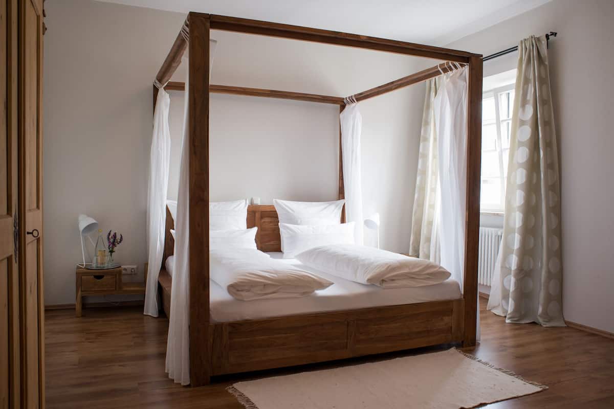A four-poster canopy bed with white linens, a bedside table with a lamp and vase, and a view of the outdoors through a window with curtains.