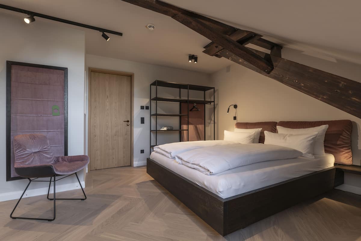 A modern bedroom with a large bed, a wooden ceiling beam, a built-in wardrobe, and a seating area with a chair.