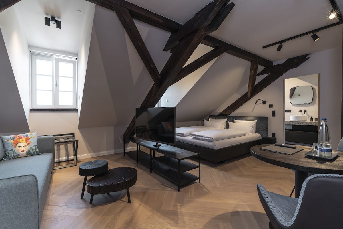 A modern loft bedroom with exposed wooden beams, a large bed, a desk, and a sofa.