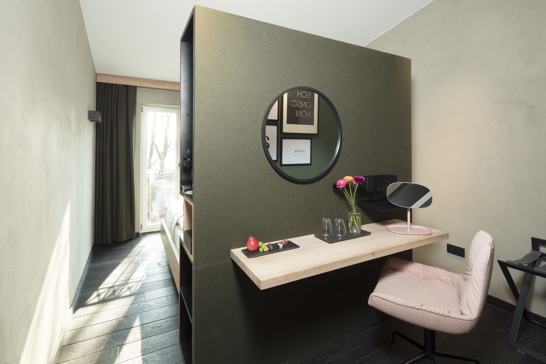 A small, modern room with a desk, chair, and a mirror. There is a window with curtains, a door, and a view of trees outside.