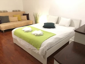 A neatly made bed with a green throw, a white headboard, and a beige sofa with pillows in a room with wooden flooring. - New Green Studio (Bucharest)