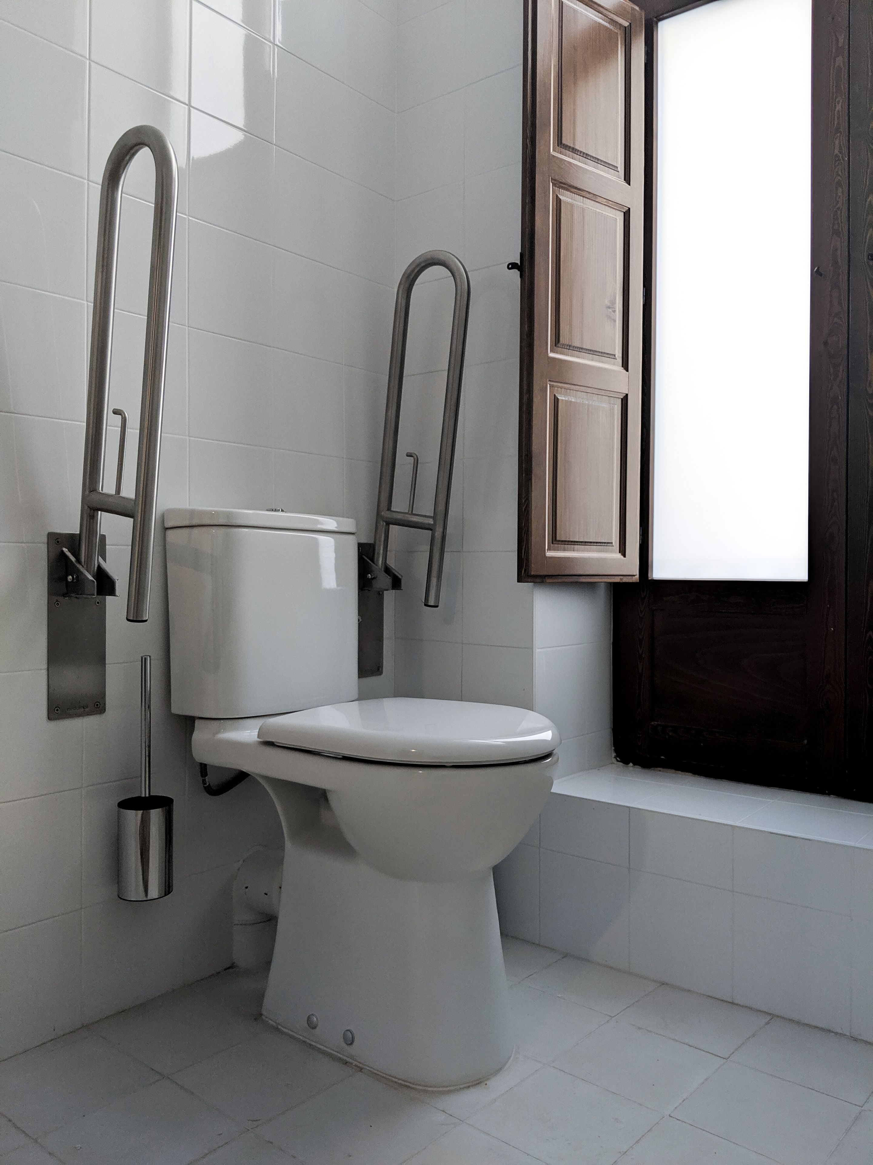 A modern bathroom with a white toilet, a grab bar, and a window with wooden shutters.