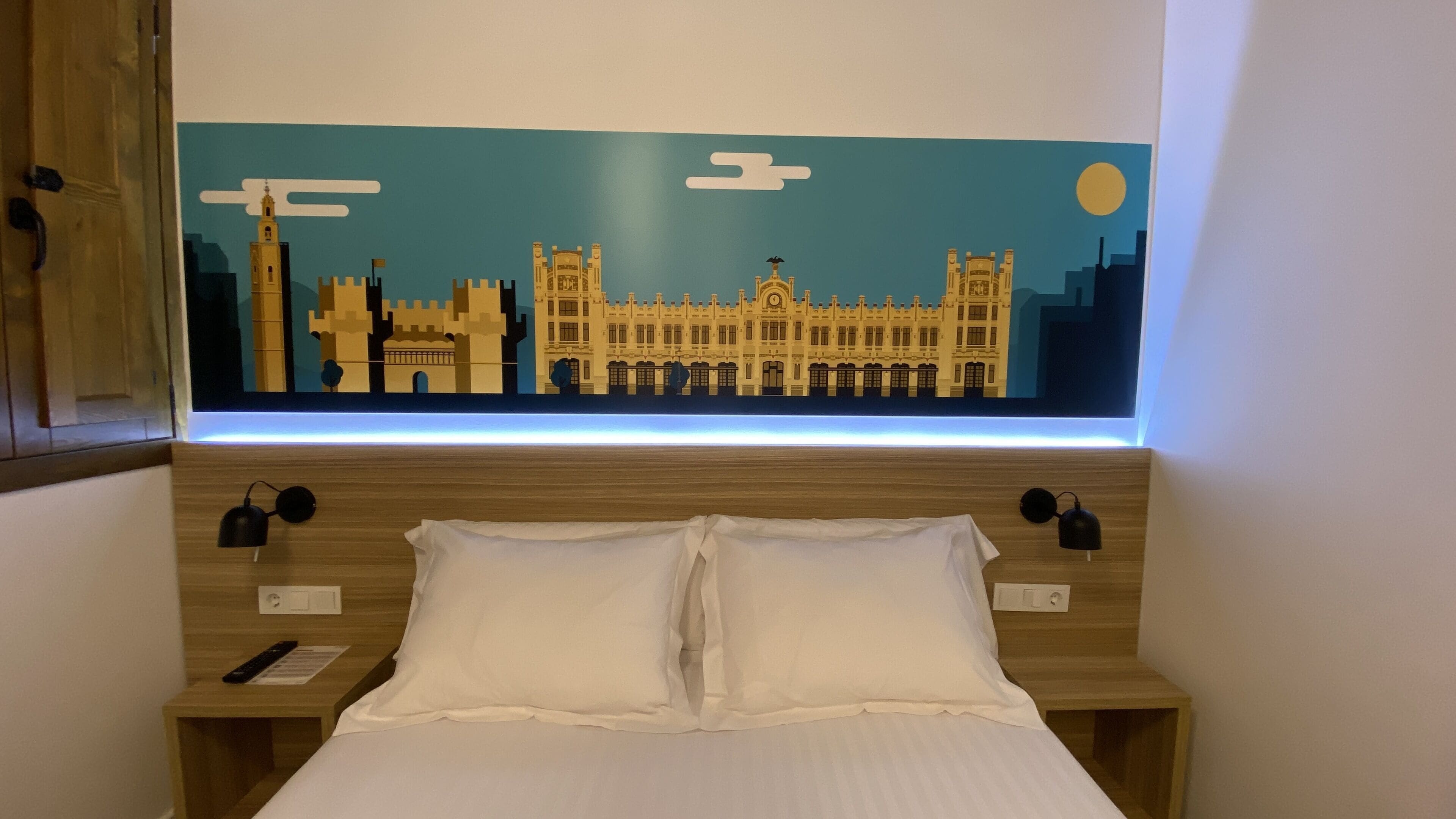 Habitación de hotel con una cama, dos mesitas de noche, una lámpara de pared y una gran obra de arte con un motivo urbano sobre la cama.