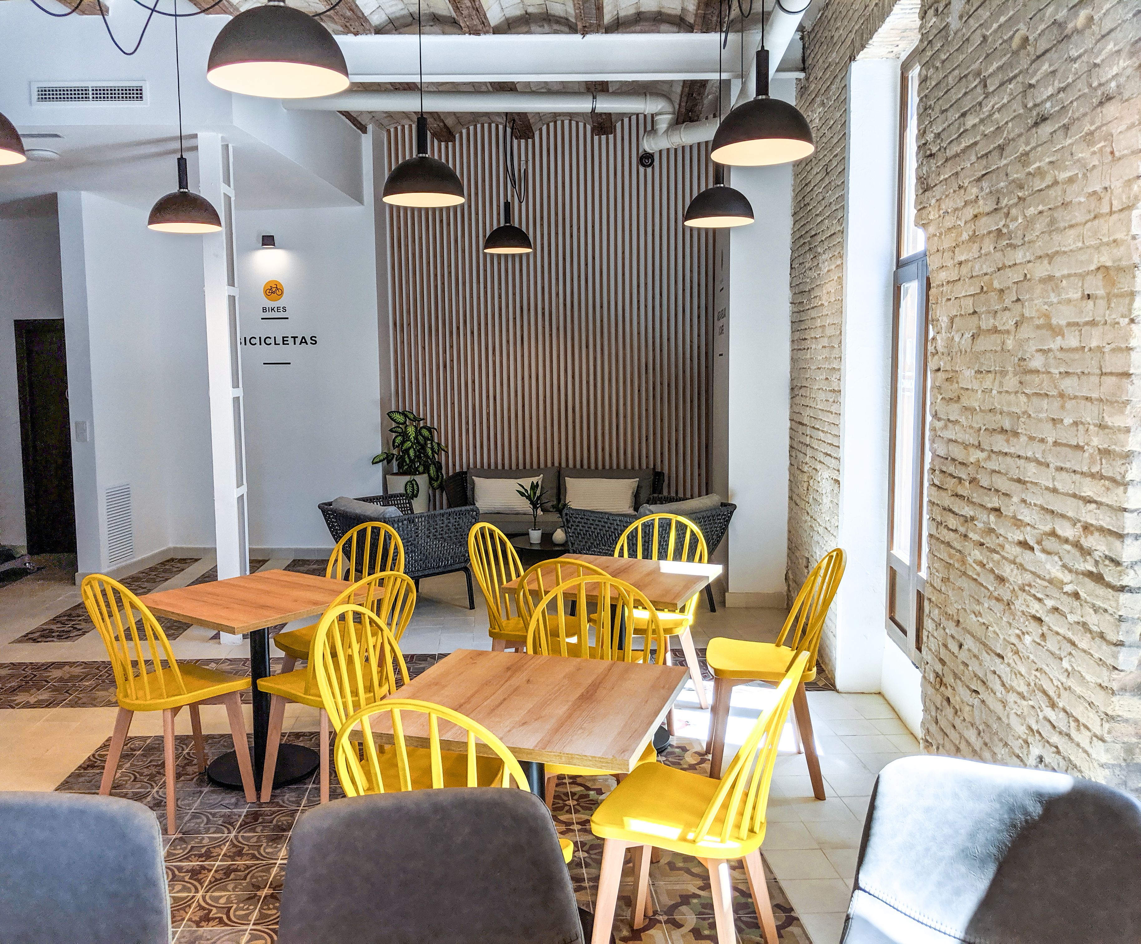 Un comedor con sillas amarillas y mesas de madera, un revestimiento de listones de madera verticales, y lámparas colgantes.