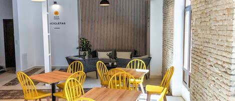Un comedor con sillas amarillas y mesas de madera, un revestimiento de listones de madera verticales, y lámparas colgantes.