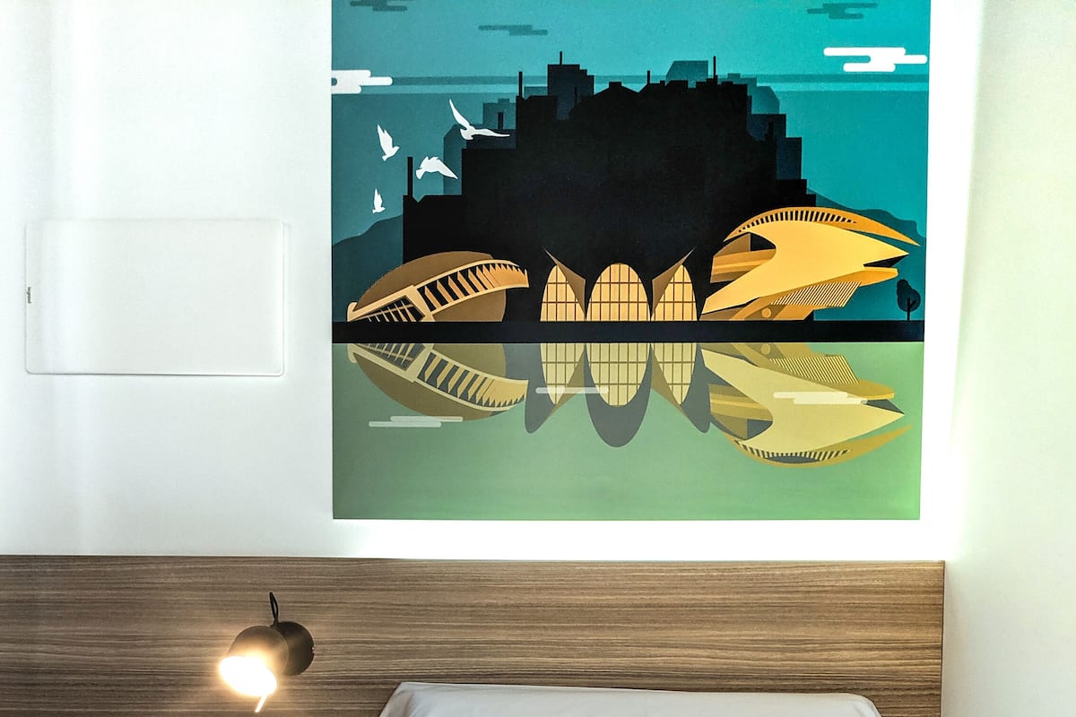 A hotel room with a bed, a nightstand with a lamp, a wall-mounted power outlet, and a cityscape artwork.